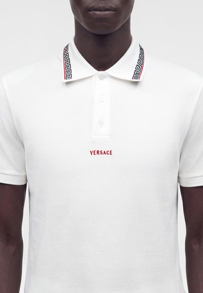 Personne à la peau foncée portant un polo blanc Versace avec un col à motif et une broderie de logo rouge sur la poitrine.