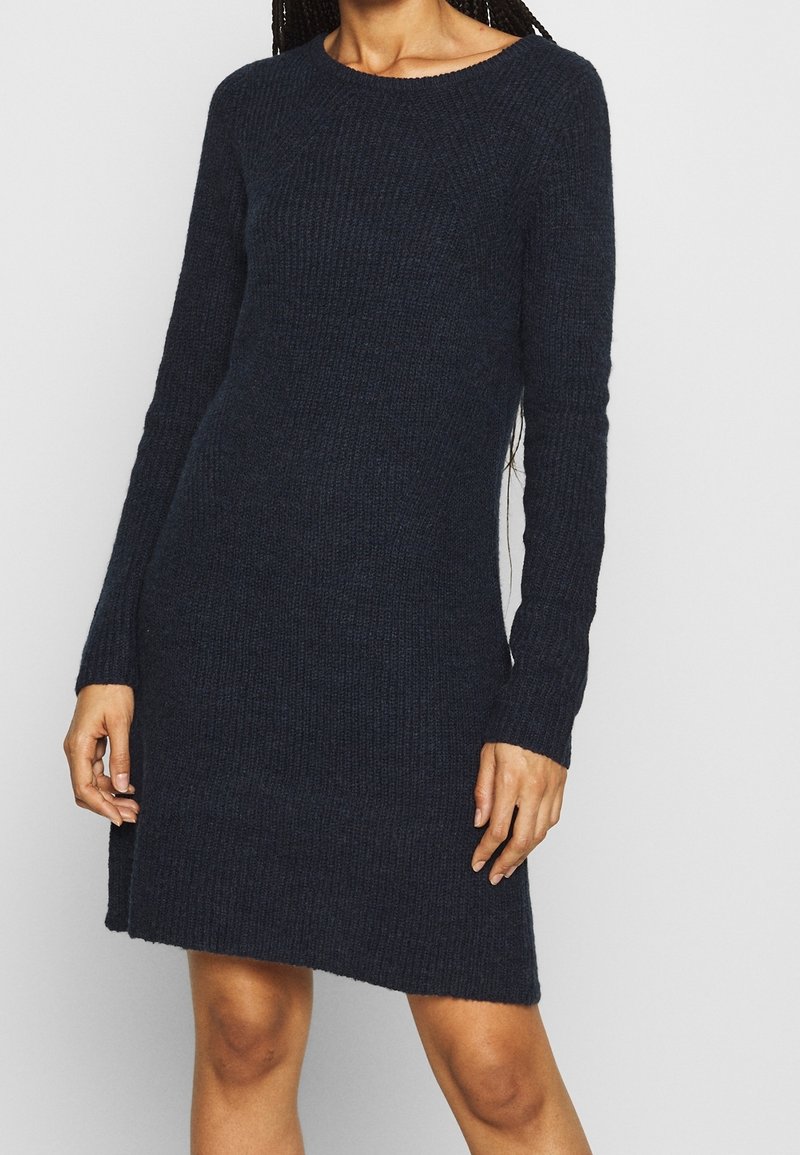 Femme portant une robe tricotée bleu marine foncé à manches longues, avec un col rond et un motif légèrement texturé.