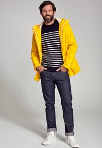 Gul regnjakke over en marineblå og hvid stribet sweater, kombineret med mørke jeans og hvide sneakers. Funktionerne inkluderer en hætte og trykknapper.