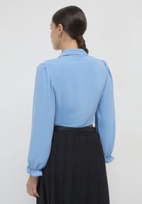 Blusa corta azzurra con maniche lunghe a sbuffo e colletto, caratterizzata da una texture liscia e una leggera lucentezza, abbinata a una gonna nera plissettata.