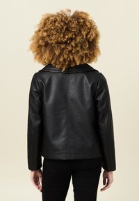 Giacca in finta pelle nera con colletto largo e cuciture verticali lungo la schiena. Indossata sopra pantaloni neri, che mettono in mostra una texture liscia.