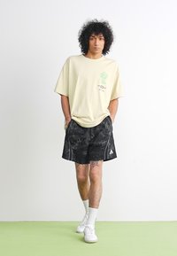 T-shirt beige chiaro con una grafica di un cactus verde, abbinato a pantaloni corti neri con motivo e dettagli bianchi, recante il logo Adidas.
