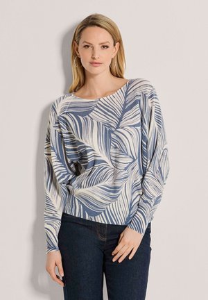 Femme aux cheveux blonds lisses, portant des boucles d'oreilles créoles et un haut à manches longues avec un motif bleu et blanc, et un jean foncé, debout contre un mur uni.