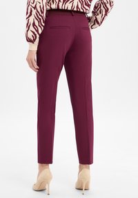 Burgundy trousers with a slim fit, made from smooth fabric, featuring two back pockets and straight hems, paired with beige high heels.