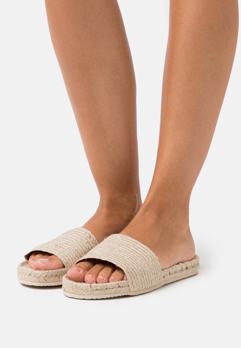 Pieds portant des sandales tissées beige à enfiler avec semelles plates sur fond blanc.
