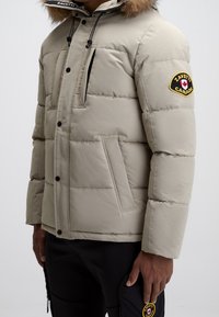 Doudoune beige avec capuche doublée de fourrure, texture matelassée, poches avant et un patch rond avec un logo en forme de feuille d'érable.