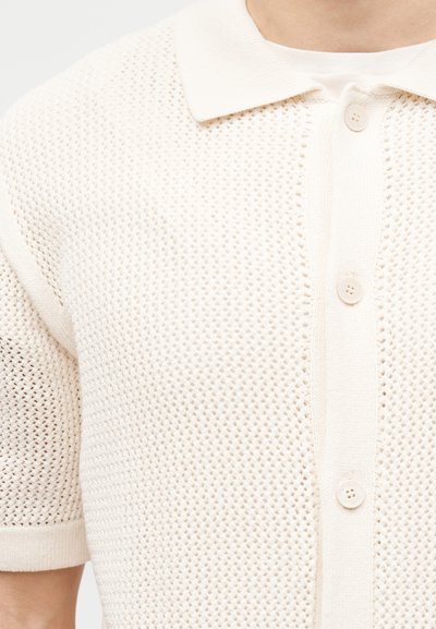 Πολό μπλούζα με κοντά μανίκια, κρεμ χρώμα, πλεκτή με υφή και ανοιχτό ύφασμα, με τρία κουμπιά μπροστά.