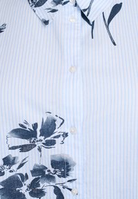 Camicia button-up di colore azzurro chiaro con righe verticali. Presenta una grande stampa floreale scura sul lato sinistro e bottoni bianchi lungo la patta.
