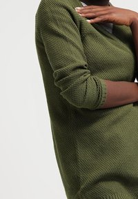 Vihreä neulepaita, jossa on tekstuuripinta, V-pääntie ja pitkät hihat. Hihansuut on käännetty, mikä korostaa rentoa istuvuutta.