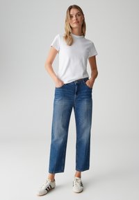 Wit T-shirt met ronde hals, blauwe jeans met rechte pijpen en vervaging op de dijen, en witte sneakers met zwarte strepen. Eenvoudige, casual outfit.