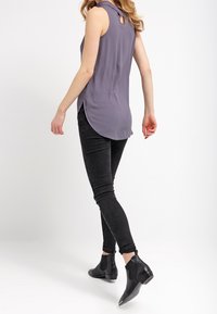 Top grigio senza maniche con dettaglio a chiave sul retro, abbinato a jeans neri aderenti e stivaletti neri su uno sfondo bianco.