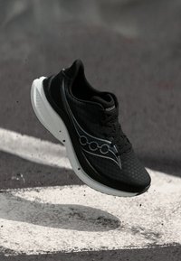 Schwarzer Sportschuh mit weißer Sohle und Logo, schwebend über einer strukturierten Asphaltoberfläche mit weißen aufgemalten Linien.