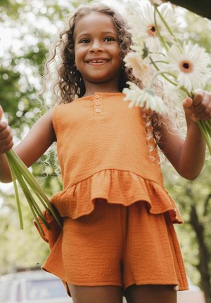 Oranje textieloutfit met een flared peplum-top en shorts. Krullend haar, met groene stelen met bloemen. Heldere natuurlijke omgeving.