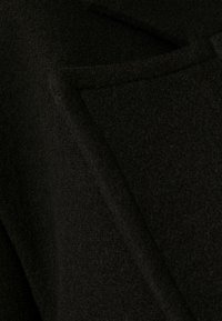 Černý vlněný kabát s texturovanou tkaninou, ostrými klopami a přední kapsou. Design je minimalistický s čistými liniemi.