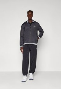 Svart jakke med glidelås, hvitstripete krage og mansjetter, kombinert med matchende bukser, vist med hvite joggesko på en ensfarget bakgrunn.