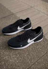 Černé sportovní boty s bílými akcenty a logem, s hladkým svrchním materiálem, zavazováním na tkaničky a polstrovanou podrážkou. Na bocích je texturovaný detail.