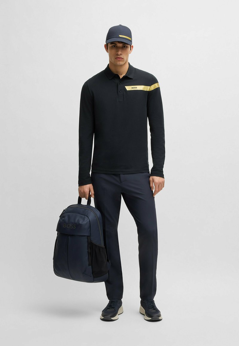 Schwarzes Langarmpolo mit gelbem Streifen, marinefarbener Kappe, marinefarbene Hose und ein marineblauer Rucksack mit einem dezenten Logodetail. Die Schuhe sind im dunklen sportlichen Stil.