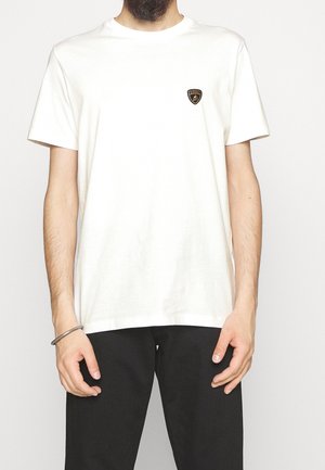 Mann trägt ein schlichtes weißes Kurzarm-T-Shirt mit einem kleinen schwarzen und goldenen Schild-Logo auf der Brust, kombiniert mit schwarzen Hosen.