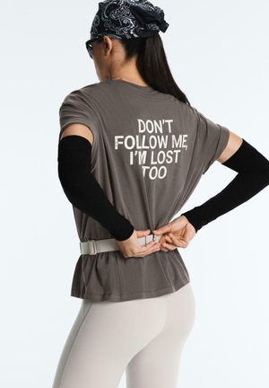 Person iført en grå skjorte med teksten "FØLG IKKE EFTER MIG, JEG ER OGSÅ VILD" på ryggen, sorte ærmer, hvidt bælte, hvide bukser og et bandana.