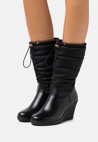 Bottes compensées noires avec un bas en cuir lisse et un dessus en tissu matelassé, dotées d'une fermeture à cordon de serrage et de détails en métal doré.