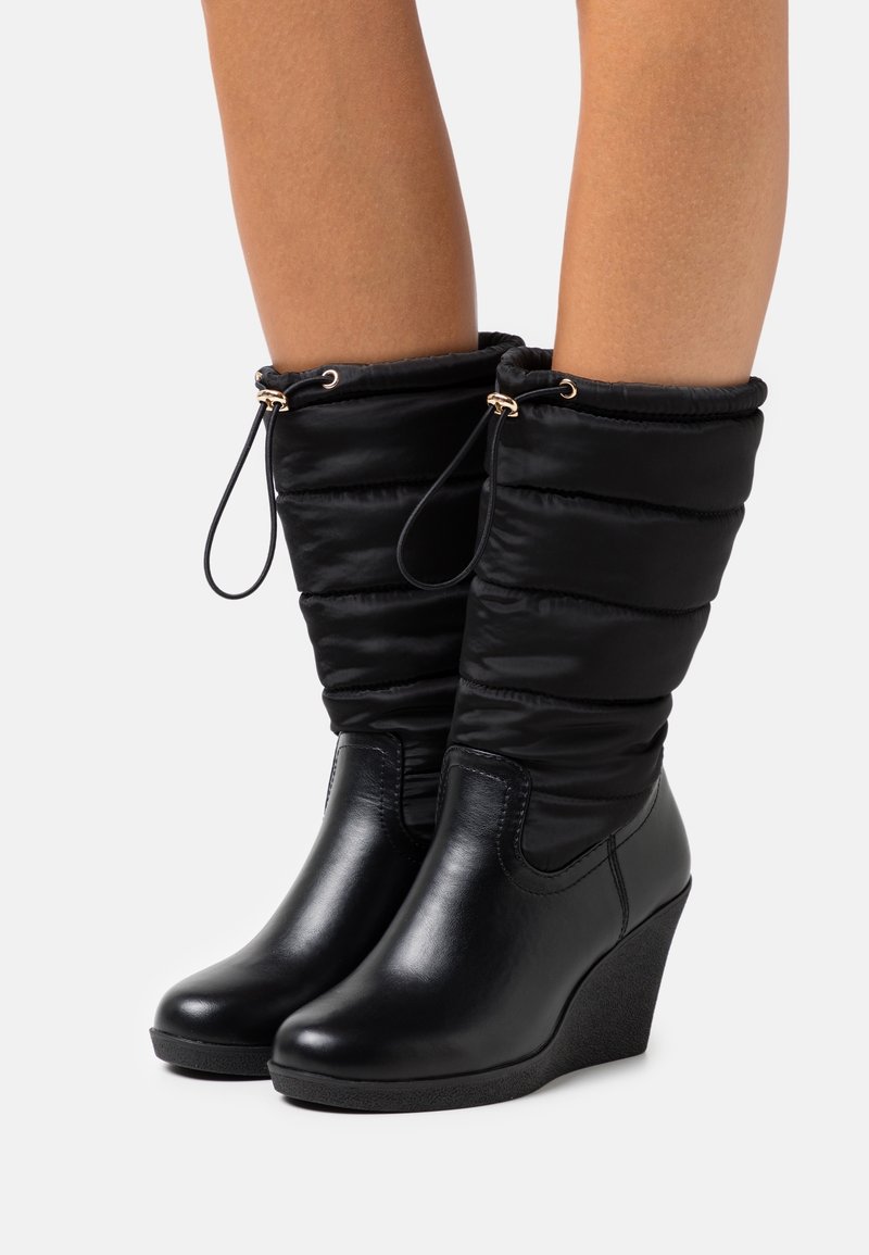 Bottes compensées noires avec un bas en cuir lisse et un dessus en tissu matelassé, dotées d'une fermeture à cordon de serrage et de détails en métal doré.