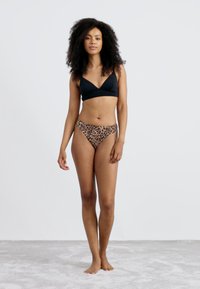 Black bikini top with a crossover design and leopard print low-rise bikini bottoms. Smooth material; model stands barefoot on a light carpet.