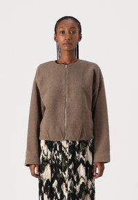 Brun fleece-jakke med glidelås foran, rund hals og avslappet passform. Har lange ermer og teksturert stoff. Mønstrede skjørt nedenfor.