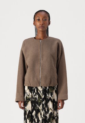 Brun fleece-jakke med glidelås foran, rund hals og avslappet passform. Har lange ermer og teksturert stoff. Mønstrede skjørt nedenfor.