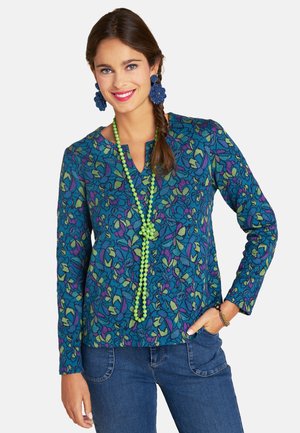 Langärmlige Bluse mit Blumenmuster in Blau, Grün und Lila, ausgestattet mit einem V-Ausschnitt und strukturiertem Stoffmuster. Kombiniert mit einer grünen Perlenkette.