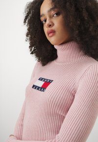 Maglione a collo alto rosa a coste con logo Tommy Jeans ricamato in navy, rosso e bianco sul petto. Design aderente.