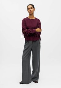 Blusa burdeos de manga larga con detalles de lazo en las mangas, combinada con pantalones de pierna ancha gris. Tejido suave, corte relajado.