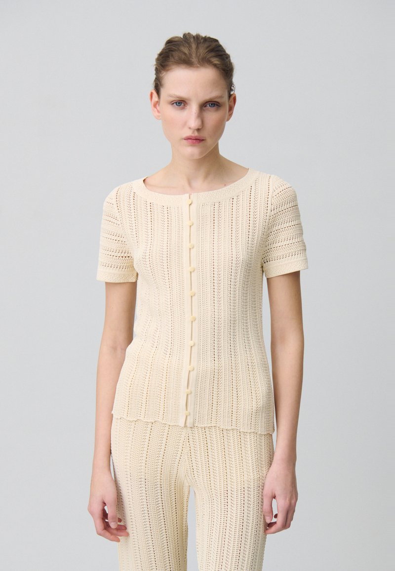 Petit haut beige en tricot avec des manches courtes, un col rond, et une fermeture à boutons. Texturé avec des motifs verticaux, un pantalon assorti visible en dessous.