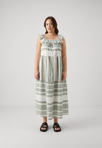 Maxikjole i hvit med grønne geometriske mønstre, rynket halslinning og ermeløs design. Kombinert med svarte sandaler med stropper.