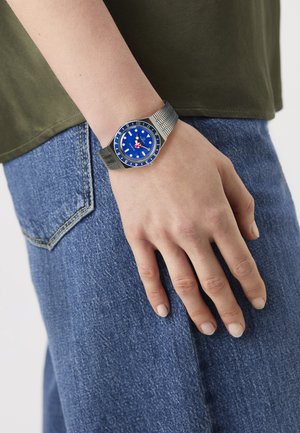 Hand wearing silver watch with blue dial showing time 10:10, resting against blue jeans and olive green shirt.