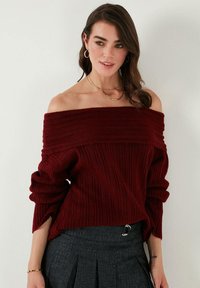 Pulover ribbed de culoare burgundy, cu umeri descoperiți, cu mâneci lungi și voloase, realizat dintr-un material texturat. Asortat cu o fustă plisată gri.