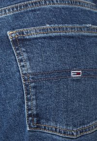 Denim jeans feature a back pocket with a red, white, and blue logo tag. The fabric is a medium blue with visible stitching and slight distressing.
