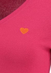 Top aderente rosa con scollo a V, realizzato in tessuto a coste. Presenta un ricamo a forma di cuore arancione sul lato sinistro.