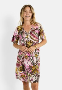 Una mujer con cabello largo y ondulado sonríe mientras lleva puesto un vestido colorido con estampado floral. Sostiene suavemente el vestido con ambas manos.