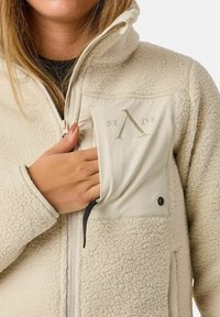 Giacca in pile beige con colletto alto, esterno testurizzato, frontale con zip e una tasca con logo in rilievo sul petto.