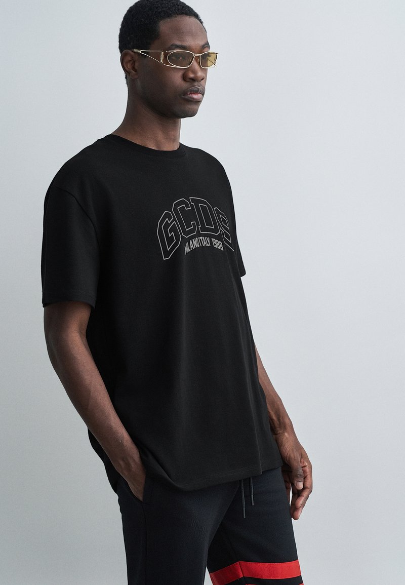 Juodas medvilninis marškinėliai su apvalia kaklo iškirpte ir trumpomis rankovėmis. Yra ryškus pilkas grafikas su „GCDS“ logotipu. Dėvimi su juodomis kelnėmis.