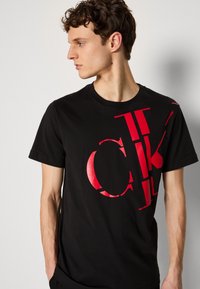 T-shirt en coton noir avec un grand motif graphique rouge, affichant des lettres en gras. Les manches courtes et le col rond complètent le design.
