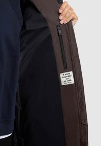 Brūna izolēta jaka, kuras iekšējā daļa rāda zip kabatu un audumu etiķeti ar tekstu. Ārējā virsma šķiet gluda; oderes krāsa ir tumša.