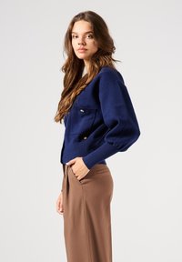 Cárdigan de punto azul marino con bolsillos frontales, botones dorados y corte holgado. Combinado con pantalones marrones de corte ajustado. Textura suave.