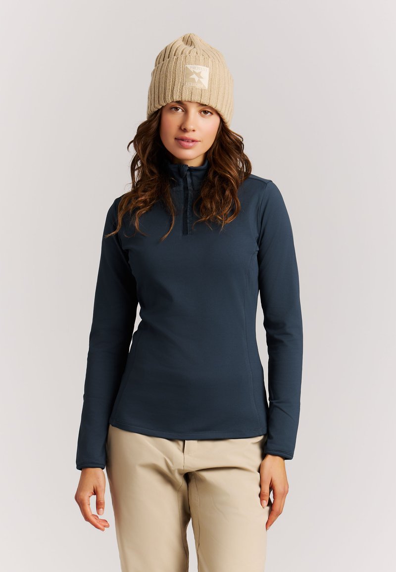 Femme portant un bonnet en laine beige, un haut zippé à manches longues bleu marine et un pantalon beige clair, debout sur fond blanc.
