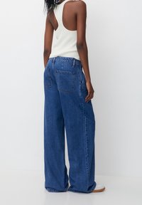 Breda blå denimjeans med hög midja och elastisk bak. Utmärkande bakfickor och en avslappnad passform, kombinerat med en ribbad vit linne.