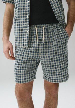 Mann trägt blau-beige karierte Shorts mit Kordelzug und ein passendes, kurzärmeliges Hemd mit Knöpfen, eine Hand in der Tasche, schwarzes Unterhemd sichtbar.