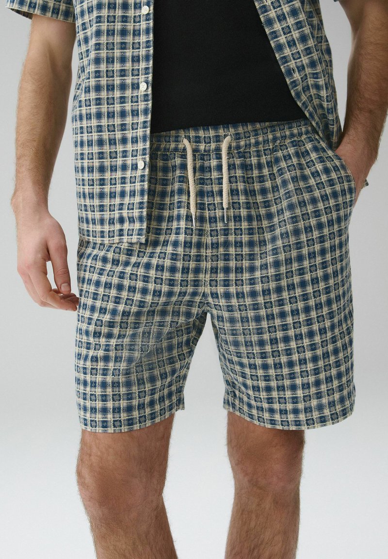 Homme portant un short à carreaux bleu et beige avec cordon de serrage et une chemise à manches courtes boutonnée assortie, main dans la poche, sous-chemise noire visible.