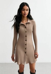 Vestido de color beige de tejido acanalado con cuello tipo camisa y mangas largas. Presenta un cierre de botones en la parte frontal y un dobladillo acampanado. Textura de tela suave.