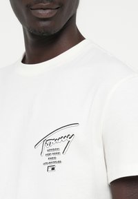 T-shirt blanc en coton avec un col rond, présentant un imprimé logo noir sur la poitrine gauche, conçu avec une texture lisse.