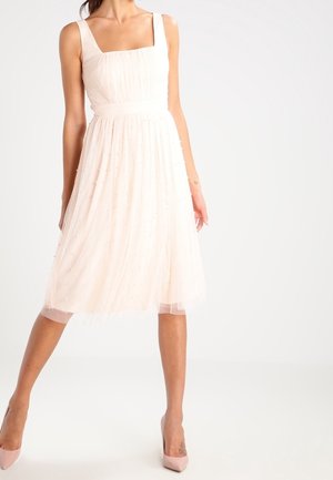 Vrouw die een mouwloze, lichtroze knielange jurk met een vierkante halslijn en roze hoge hakken draagt, staand met één voet naar voren.